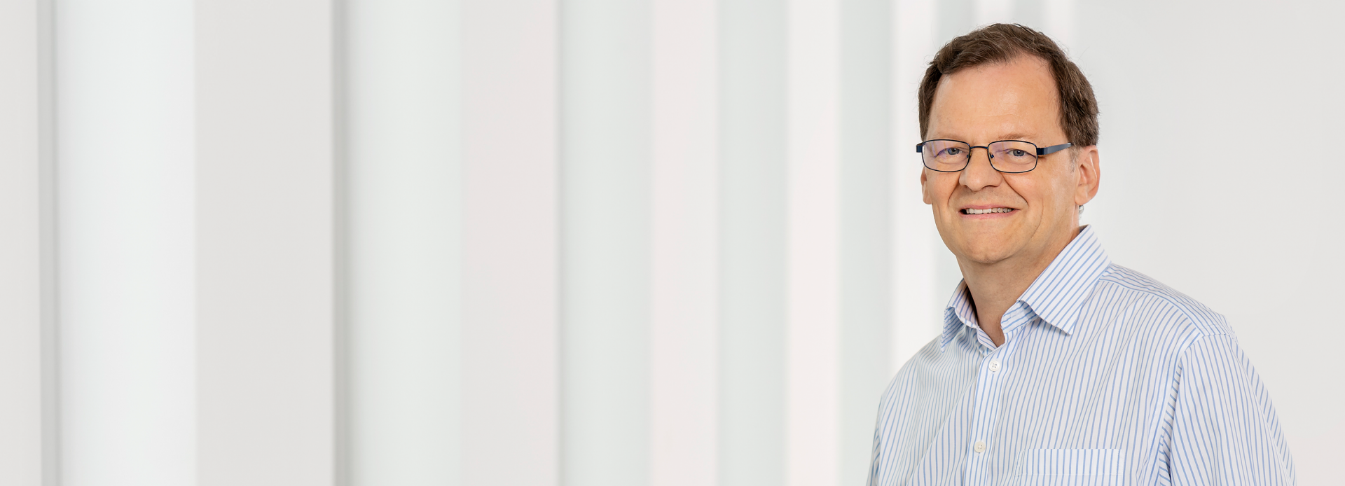 Die Person mit einem gestreiften hellblauen Hemd vor einem Hintergrund aus weißen vertikalen Linien oder Säulen.