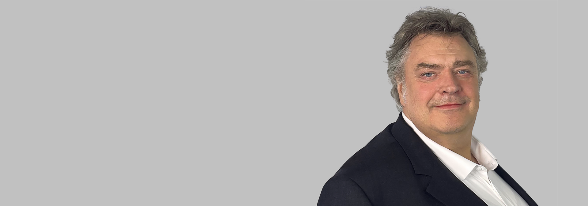 Das Bild zeigt eine Person, die formell gekleidet ist, möglicherweise mit einem Anzug und weißem Hemd, vor einem grauen Hintergrund. Es scheint sich um ein Portrait zu handeln.